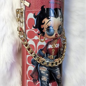 May include: A red and white tumbler featuring a Betty Boop illustration in a stylish outfit, with the text "BETTY BOOP COACH NEW YORK". The tumbler has a gold chain handle and a zipper design.