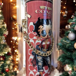 May include: A red cylindrical tumbler with a Betty Boop design and the text "Betty Boop Coach New York". It has a gold chain handle and a zip closure. The design features a Betty Boop illustration in a red top and ripped jeans.