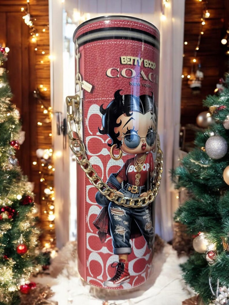 May include: A red cylindrical tumbler with a Betty Boop design and the text "Betty Boop Coach New York". It has a gold chain handle and a zip closure. The design features a Betty Boop illustration in a red top and ripped jeans.
