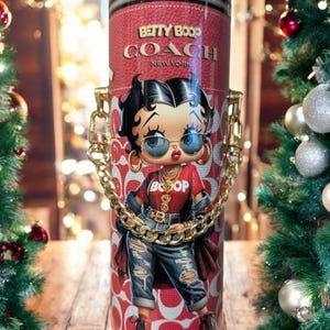 May include: A red and white Coach tumbler with a Betty Boop design. It has a gold chain handle and the words "Betty Boop Coach New York" are printed on the front. The background is blurred with Christmas decorations.