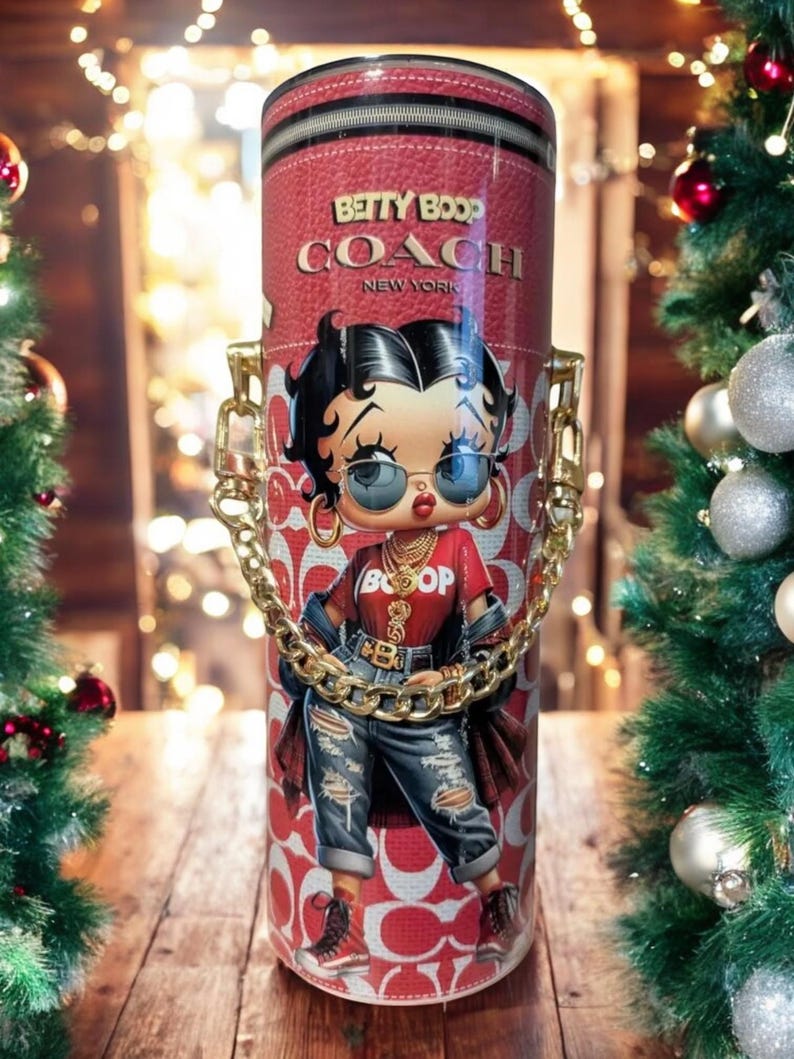 May include: A red and white Coach tumbler with a Betty Boop design. It has a gold chain handle and the words "Betty Boop Coach New York" are printed on the front. The background is blurred with Christmas decorations.