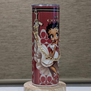 May include: A red tumbler with a Betty Boop design in a white dress, a gold chain, and the Coach logo. The cylindrical tumbler sits on a wooden stand. The design incorporates the Coach logo pattern.