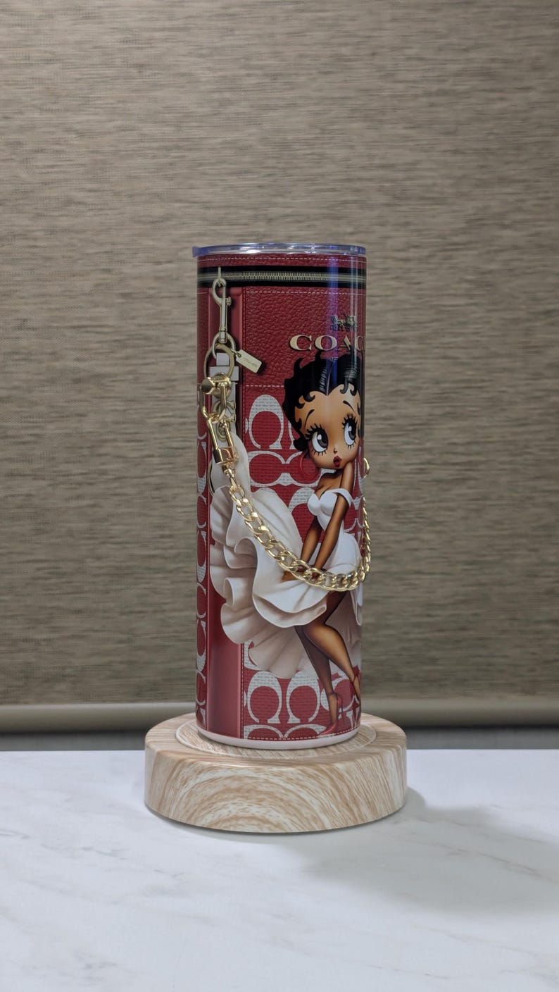 May include: A red tumbler with a Betty Boop design in a white dress, a gold chain, and the Coach logo. The cylindrical tumbler sits on a wooden stand. The design incorporates the Coach logo pattern.