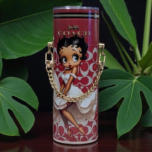 May include: Red Coach tumbler with a Betty Boop design. It has a gold chain handle and a red and white pattern. The Coach logo is at the top. The cylindrical tumbler is likely made of metal. The design features a cartoon character.