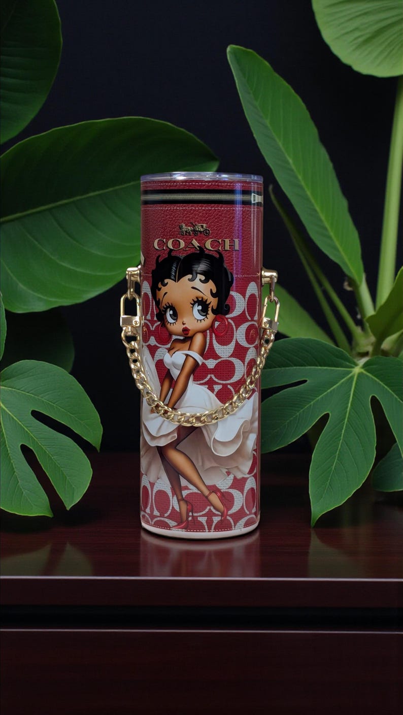 May include: Red Coach tumbler with a Betty Boop design. It has a gold chain handle and a red and white pattern. The Coach logo is at the top. The cylindrical tumbler is likely made of metal. The design features a cartoon character.