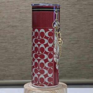 May include: A red and white tumbler with a repeating pattern of interlocking Cs. The tumbler has a clear lid and a gold-coloured chain with a charm. The tumbler is cylindrical and sits on a wooden base.