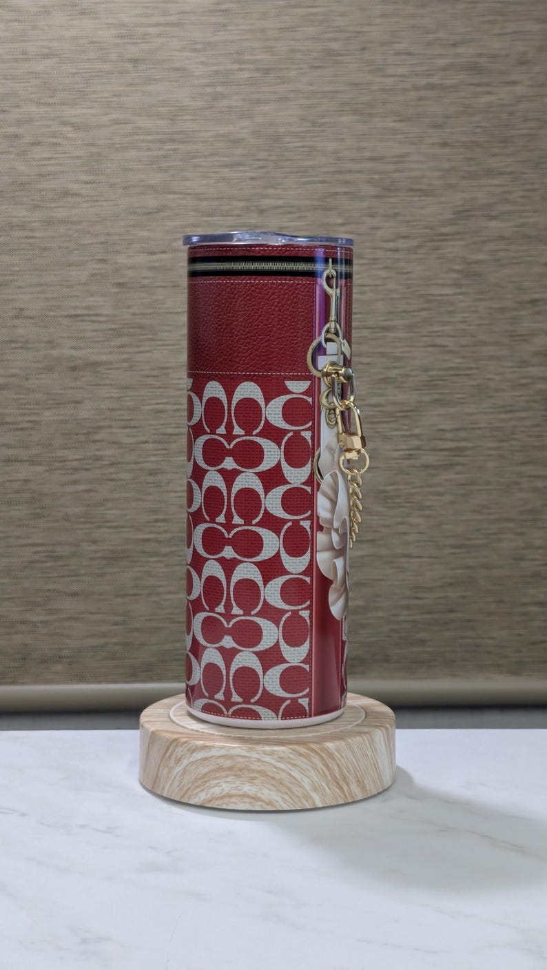 May include: A red and white tumbler with a repeating pattern of interlocking Cs. The tumbler has a clear lid and a gold-coloured chain with a charm. The tumbler is cylindrical and sits on a wooden base.