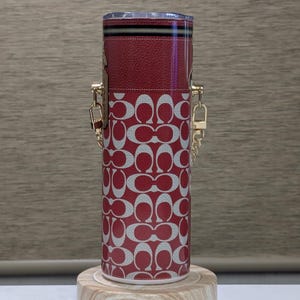 May include: A cylindrical red and white patterned tumbler with gold-coloured chain handles. The tumbler has a red leather top with a zip and a clear lid. It is displayed on a small wooden stand.