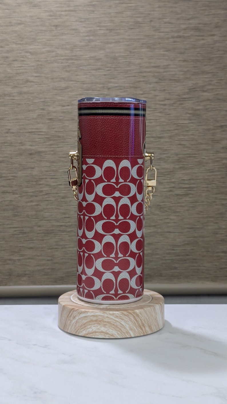 May include: A cylindrical red and white patterned tumbler with gold-coloured chain handles. The tumbler has a red leather top with a zip and a clear lid. It is displayed on a small wooden stand.