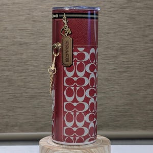 May include: Red and white Coach tumbler with a repeating logo pattern. The tumbler has a zipper detail at the top and a gold-tone charm with the Coach logo. It is displayed on a wooden stand.