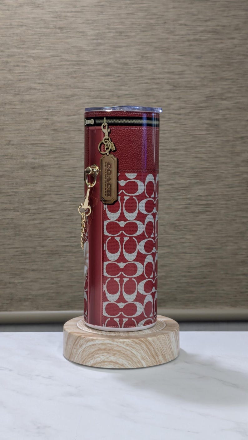 May include: Red and white Coach tumbler with a repeating logo pattern. The tumbler has a zipper detail at the top and a gold-tone charm with the Coach logo. It is displayed on a wooden stand.