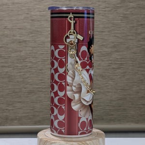 May include: A red and white tumbler with a zip design and a gold keyring. The tumbler features a repeating pattern of interlocking "C" shapes and a graphic of a woman. The tumbler is displayed on a wooden stand.