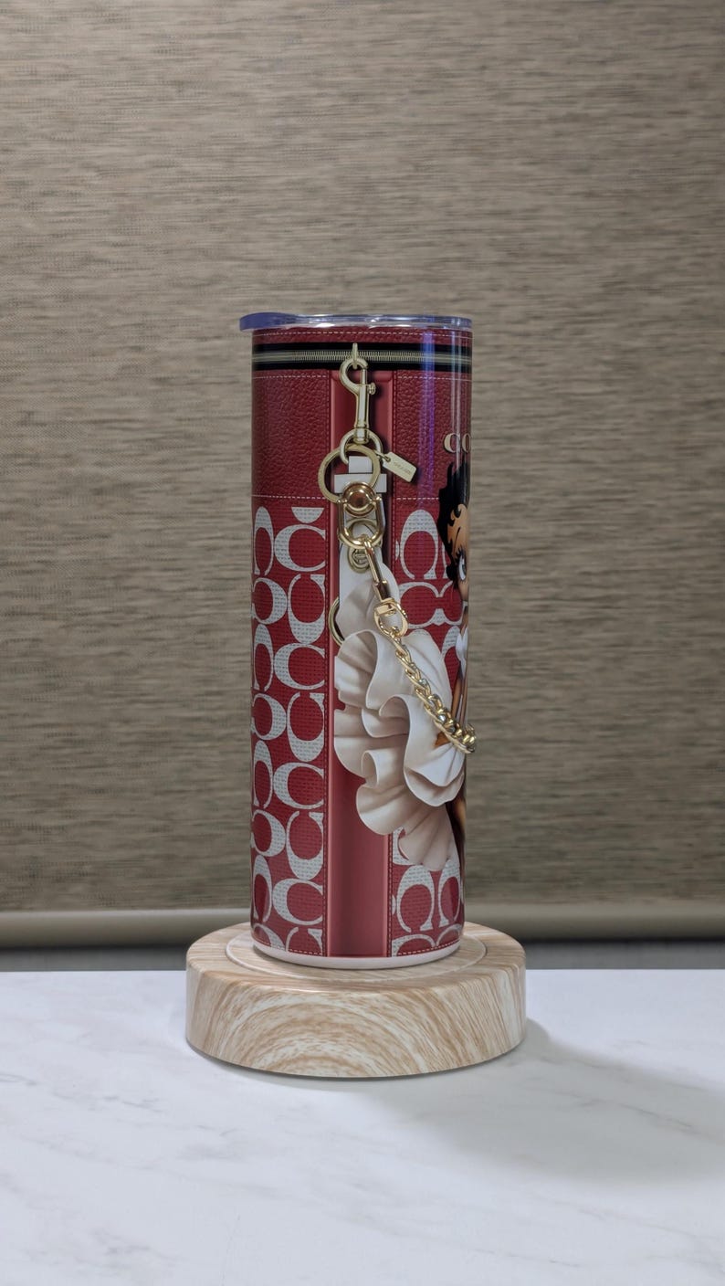May include: A red and white tumbler with a zip design and a gold keyring. The tumbler features a repeating pattern of interlocking "C" shapes and a graphic of a woman. The tumbler is displayed on a wooden stand.