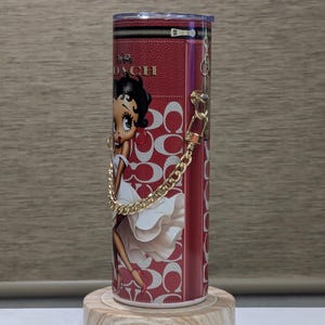 May include: A red and white tumbler featuring a Betty Boop illustration and the Coach logo. The tumbler has a gold chain handle and is displayed on a wooden stand. The design includes the Coach logo pattern.