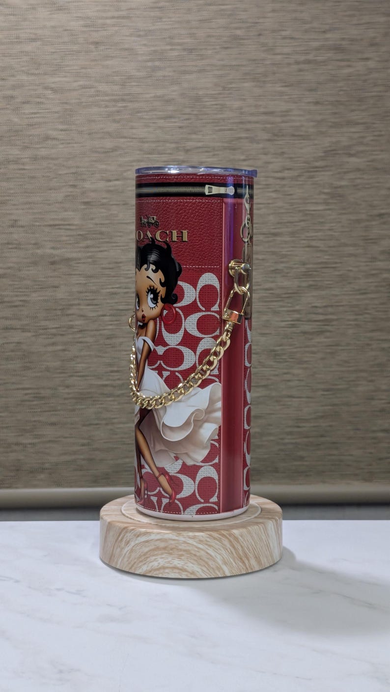 May include: A red and white tumbler featuring a Betty Boop illustration and the Coach logo. The tumbler has a gold chain handle and is displayed on a wooden stand. The design includes the Coach logo pattern.