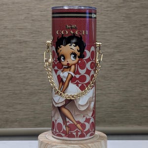 May include: Red Coach tumbler with a Betty Boop graphic. It has a gold chain handle and a red and white pattern. The Coach logo is at the top. The tumbler is displayed on a wooden stand.