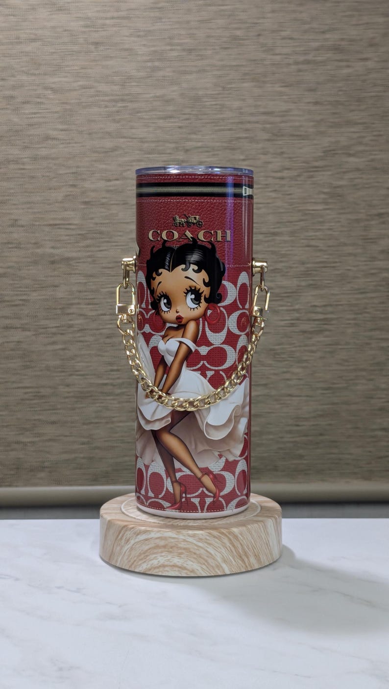 May include: Red Coach tumbler with a Betty Boop graphic. It has a gold chain handle and a red and white pattern. The Coach logo is at the top. The tumbler is displayed on a wooden stand.