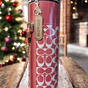 May include: A red and white Coach-branded tumbler with a key chain and a leather tag. The tumbler features a repeating pattern of the Coach logo and a faux leather design. The tumbler is cylindrical and has a black rim.