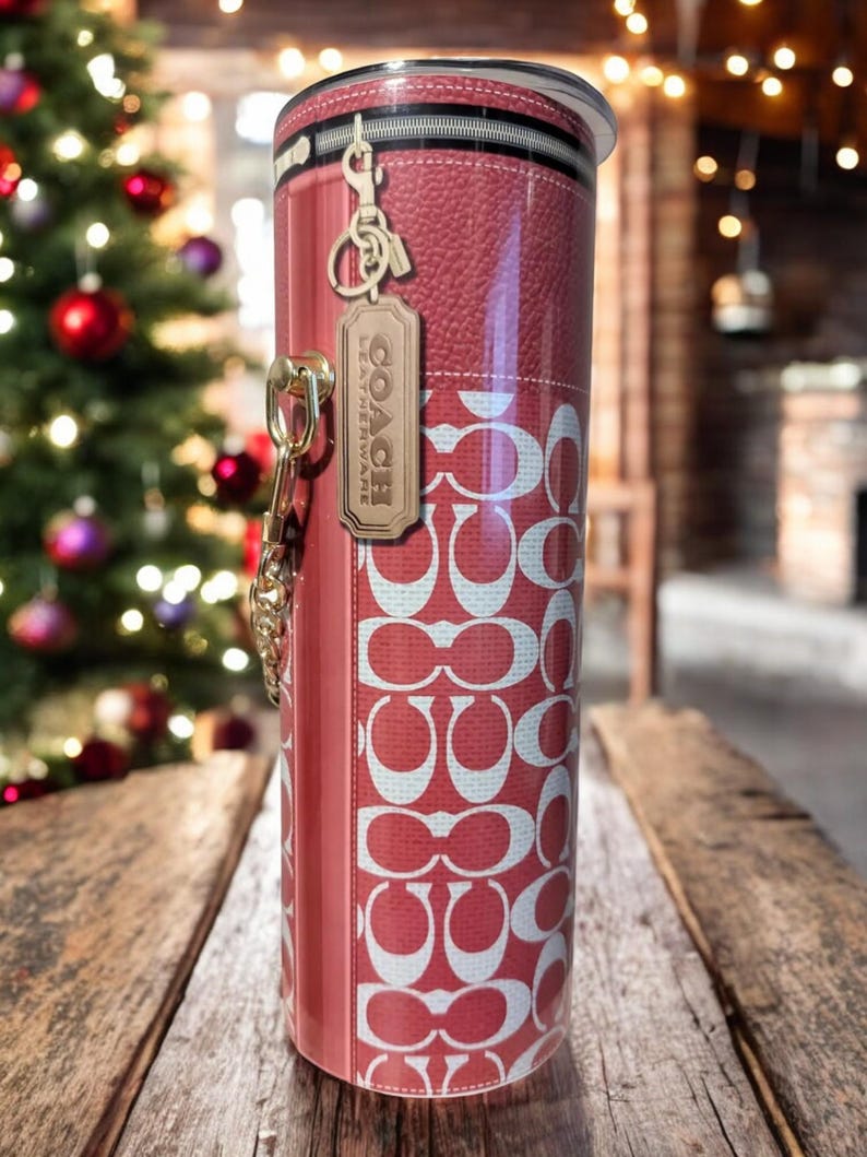 May include: A red and white Coach-branded tumbler with a key chain and a leather tag. The tumbler features a repeating pattern of the Coach logo and a faux leather design. The tumbler is cylindrical and has a black rim.