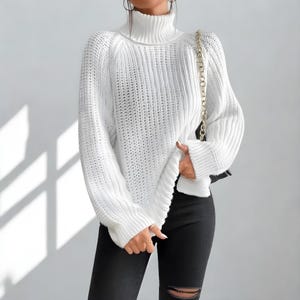 Può includere: Un maglione bianco a collo alto in maglia grossa con maniche lunghe. Il maglione ha una vestibilità ampia e una trama a coste. La modella indossa jeans neri. È visibile una borsa con tracolla a catena dorata.