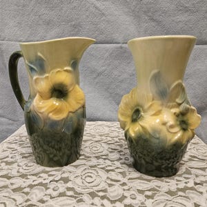 May include: Two ceramic vases with floral designs. One is a pitcher with a handle, the other a vase. Both have a cream-colored base with yellow and green floral accents. The pitcher is on the left, and the vase is on the right.