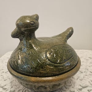 May include: A ceramic lidded dish shaped like a duck with a mottled olive green glaze. The lid features carved feather details and a sculpted duck head, while the base is a simple bowl with a textured, earthy brown and green finish.