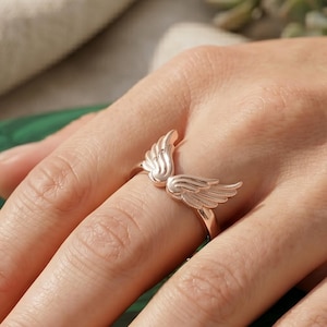 May include: A rose gold ring featuring a detailed wing design. The ring is displayed on a hand, showcasing the delicate feather details. The ring is a piece of jewelry, perfect for adding a touch of elegance.