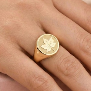 May include: A gold signet ring featuring a detailed maple leaf design. The ring has a polished finish and a round face with the leaf emblem. The ring is shown on a hand, highlighting the jewelry's design and style.