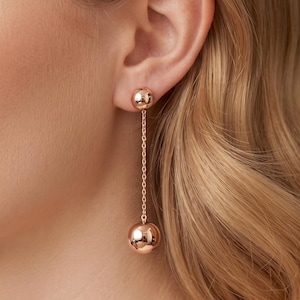 May include: Rose gold drop earrings featuring a small sphere stud and a larger sphere suspended by a delicate chain. The earrings are shown on a person with blonde hair, offering a view of the ear and the jewelry's design.