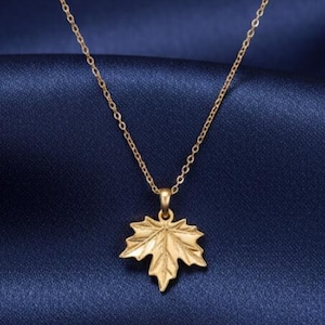 May include: A gold-coloured necklace featuring a maple leaf pendant. The pendant, with detailed leaf veins, hangs from a delicate chain. The background is a dark blue fabric.