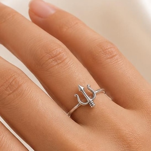 May include: A silver-toned ring featuring a small trident symbol as the centerpiece. The ring has a thin, polished band and is worn on the ring finger. The trident design is centered on the band, showcasing a three-pronged shape with a small horizontal bar detail at the base of the prongs.