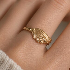 May include: A gold ring featuring a fan-shaped design. The ring is worn on a finger, with the fan detail radiating outwards. The ring's design is set against a soft, neutral background, highlighting the jewelry's elegant simplicity.
