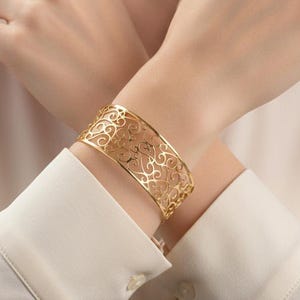 May include: A gold-colored cuff bracelet with an openwork filigree design. The bracelet is wide and wraps around the wrist. The intricate pattern features swirling, floral-like shapes. The bracelet is worn on a person's wrist, against a white long-sleeved shirt.