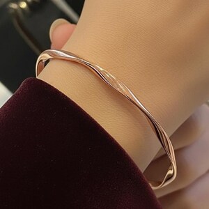 Può includere: Un bracciale in metallo ritorto color oro rosa. Il bracciale ha un design liscio e fluido, con un sottile motivo a onde. Il gioiello è indossato su un polso e si vede la manica di un capo bordeaux.