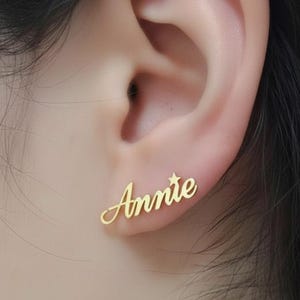 Könnte beinhalten: Goldfarbene Ohrstecker mit dem Namen "Annie" in Schreibschrift und einem kleinen Stern. Die Ohrringe werden an einem Ohr getragen, um das Design und die Passform zu zeigen.