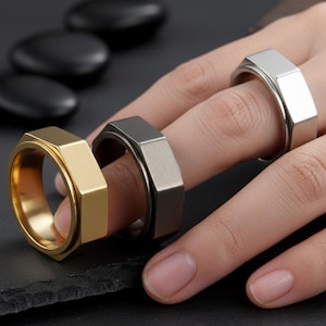 May include: Three metal rings in gold, silver, and gunmetal gray are displayed. The rings have a hexagonal design. One ring is on a finger, while the other two are next to it. The background is blurred, with black stones.