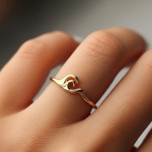 Può includere: Un anello dorato con un design a onda. L'anello presenta un design delicato, con la forma dell'onda come elemento centrale. Il gioiello è indossato su un dito, evidenziando il suo stile elegante e minimalista.