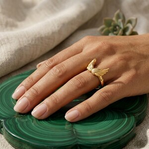 May include: A gold-colored ring featuring a wing design. The ring is worn on a hand resting on a green, circular surface. The ring's wings are detailed and spread out, creating a delicate and elegant look. The ring is a piece of jewelry.