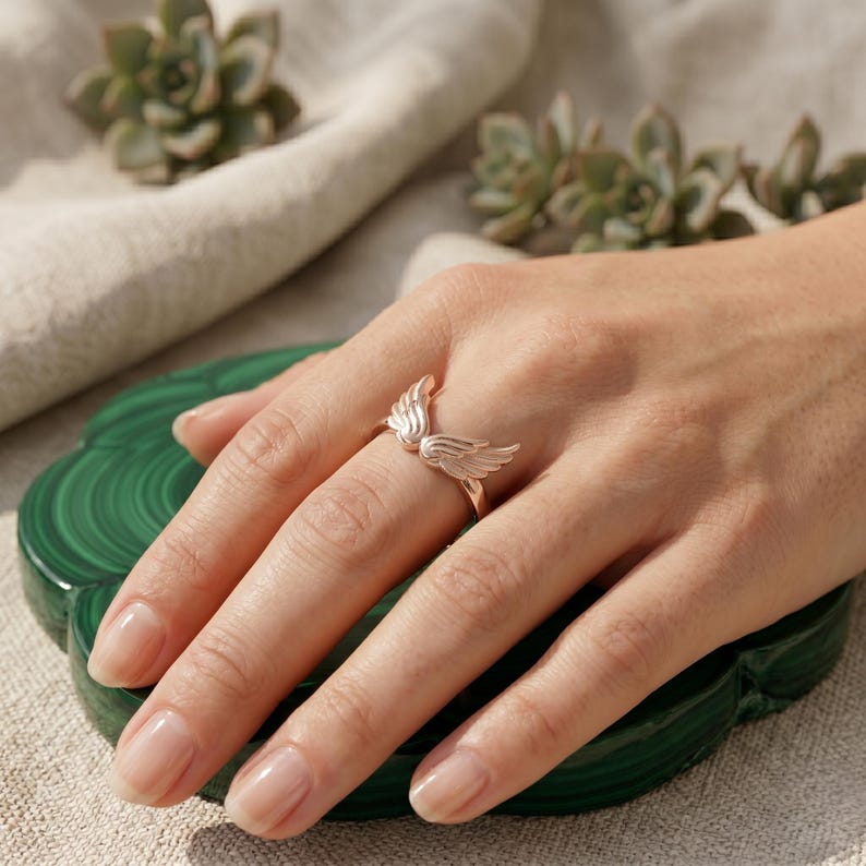 May include: A rose gold ring featuring a detailed wing design. The ring is displayed on a hand, showcasing the delicate feather details. The ring is a piece of jewelry, perfect for adding a touch of elegance.