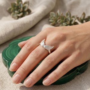 May include: A silver ring featuring a pair of detailed angel wings. The ring is worn on a finger, showcasing the delicate design. The wings are spread out, creating a unique and elegant look. The ring is set against a soft background.