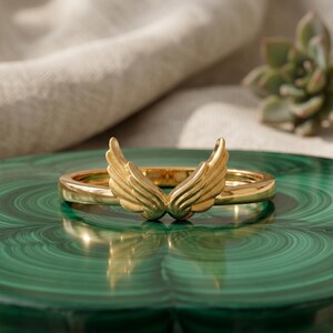 May include: A gold-colored ring with a wing design. The ring is set on a green surface, with a succulent plant in the background. The wings are detailed and appear to be made of metal.