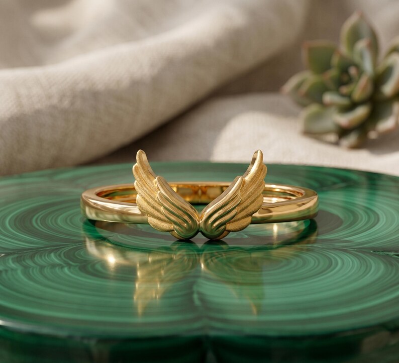 May include: A gold-colored ring with a wing design. The ring is set on a green surface, with a succulent plant in the background. The wings are detailed and appear to be made of metal.