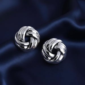 Può includere: Un paio di orecchini a bottone argentati a forma di nodo. Gli orecchini sono rotondi con un design intrecciato, che riflette la luce. Sono appoggiati su un tessuto blu scuro e setoso, creando un contrasto.