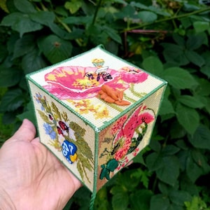 以下が含まれることがあります： 花柄刺繍が施された立方体の手作りボックス。大きなピンクのポピー、ミツバチ、その他のカラフルな花が特徴です。縁は緑色でトリミングされ、緑色のタッセルが下からぶら下がっています。小さなオレンジ色の粘土のフィギュアが上に置かれています。