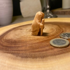 Könnte beinhalten: Kleine, hellbraune Holzfigur einer Person in einer Kapuzenrobe, die einen Stab hält. Die Figur steht auf einer kreisförmigen Holzoberfläche, mit zwei Münzen in der Nähe. Die Holzmaserung ist sichtbar.