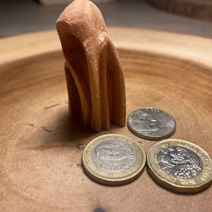 Puede incluir: Una escultura abstracta de color marrón claro con forma de capucha se encuentra sobre una superficie de madera. Tres monedas, incluyendo una moneda de euro y otras dos monedas, se colocan cerca de la escultura. La madera tiene una veta natural.