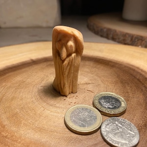 Op de afbeelding: Een klein, gesneden houten beeldje van een figuur met een mantel, staand op een houten oppervlak. Drie munten zijn bij het beeldje geplaatst. Het hout heeft een natuurlijke, lichtbruine kleur.