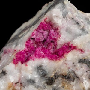 Puede incluir: Primer plano de un espécimen mineral natural y rugoso. La piedra presenta una mezcla de tonos blancos, grises y negros, con una vibrante bolsa de cristales de color rosa brillante. La textura es áspera y desigual, con estructuras cristalinas visibles.