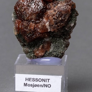 Puede incluir: Un espécimen de piedra preciosa de hessonita marrón rojizo rugoso sobre un pequeño expositor transparente. La piedra tiene una estructura cristalina y está colocada sobre una matriz verde oscuro. La etiqueta dice "HESSONIT Mosjøen/NO".