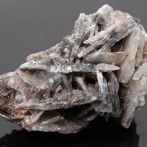 Puede incluir: Un espécimen mineral en bruto que presenta un grupo de cristales translúcidos en forma de hoja en tonos blanco, gris y marrón. La estructura cristalina muestra formaciones afiladas y alargadas con una textura natural y sin pulir.